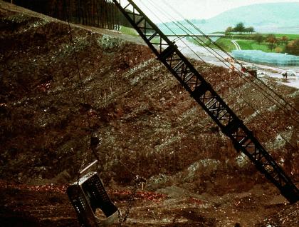 Bergwärts einfallende Schichtung der Großschollenrutschung innerhalb der Grabfeld-Formation, während Sanierungsarbeiten aufgenommen Das Bild zeigt eine Grube. Im Vordergrund verdeckt ein Kran teilweise das Sichtfeld. Im Hintergrund Felder mit einzelnen Obstbäumen.