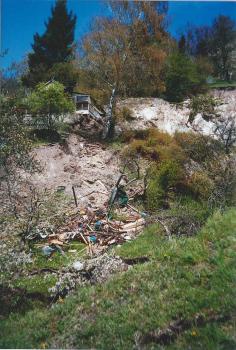 Zerstörte Garteneinrichtungen am Rand der Rutschung im Kirchsteigtobel bei Urbach Das Bild zeigt im Vordergrund durch eine Rutschung zerstörte Garteneinrichtungen auf einer nach links abfallenden Wiese. Im Mittelgrund ist die Rutschung zu sehen, im Hintergrund und weiter oben am Hang eine Hütte und Bäume.