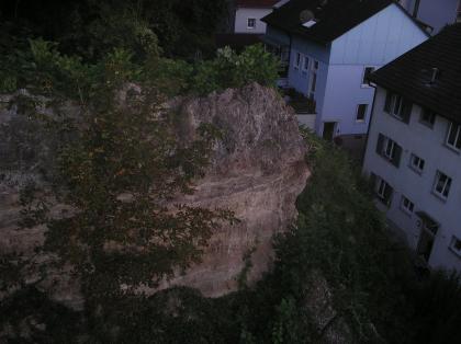 Blick von oben auf die Ausbruchstelle Blick von oben auf einen mit Pflanzen bewachsenen Felshang, von dem ein großes Stück abgebrochen ist. Rechts am Bildrand befinden sich Wohnhäuser.