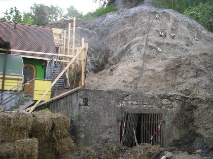 Aufbringung des Steinschlagschutznetzes Im Mittelgrund des Bildes befindet sich ein Felshang, welcher durch ein Netz gegen Steinschlag gesichert wird. In der linken Bildhälfte befindet sich ein abgesichertes Haus.