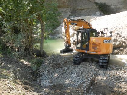 Bagger beim Beräumen des Bachbettes Im Hintergrund des Bildes befindet sich eine Felswand, vor der ein großer Felsblock in einem Gewässer liegt. Vor dem Felsblock steht ein Bagger halb im Wasser, der mit seiner Schaufel im Gewässer gräbt. Am linken Bildrand stehen Bäume.