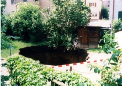 Das Bild zeigt einen kreisförmigen Krater im Garten eines Wohngebiets. Am hinteren Rand des Erdfalls stehen noch Bäume. Der Bereich ist abgesperrt.