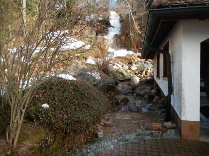 Gebäude- und Sachschäden sowie abgelagerte Geröllfracht auf dem südlich an den Oberen Großbach angrenzenden Grundstück Nr. 8 am Hangfuß Am rechten Bildrand ist im Vordergrund ein Haus zu sehen. Hinter und neben dem Gebäude und auch direkt an dessen linker Außenwand liegen einige große Steine. Wasser fließt den Hang hinter dem Gebäude hinab und gelangt am Haus vorbei bis vor den Eingang.