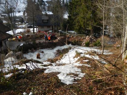 Beseitigung der Schäden in der bebauten Ortslage von Menzenschwand-Hinterdorf Das Bild zeigt einen stark angeschwollenen Bach, der an einem Gebäude entlang auf eine Brücke zufließt. Die Brücke ist mit Schlamm, Steinen und Ästen bedeckt. Dahinter stehen zwei Bagger und einige Personen auf einer Straße; im Hintergrund weitere Häuser.