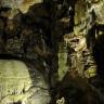 Blick in das Innere einer Höhle. Teile der Höhle - etwa links vorne ein geglätteter Felsbuckel mit Tropfsteinbildungen oder rechts hinten eine zerklüftete Wand - sind beleuchtet und zeigen gelblich graue Farben.