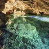 Blick in das Innere einer Höhle. Der blaugrün schimmernde Boden wird von einem See bedeckt. Die zerklüftete Decke ist gewölbt und hellbraun gefärbt, wo Licht sie erhellt.