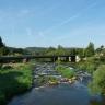 Blick auf einen Flusslauf, der zum Betrachter hin über mehrere flache Stufen hinabfließt. Eine schmale Eisenbahnbrücke überquert den Fluss.