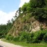 Blick von der Straße, die sich im linken Vordergrund befindet, auf eine steile, teilweise von Sträuchern und Büschen überwachsene Felswand.