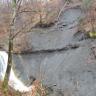 Blick auf eine steile graue, teilweise bewachsene Gesteinswand. Links schäumt ein schmaler Wasserfall. Davor steht auf einem Felsvorsprung ein hoher, schlanker Baum.
