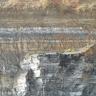 Das Bild zeigt mehrere Schichten eines Steinbruchs. Während rechts unten grob gebankte, grauweiße Blöcke zu sehen sind, zeigt die übrige Wand feinere Streifen. Die Farben wechseln hier von grau und weiß unten und mittig zu gelb- bis rotbraun oben.