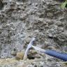 Nahaufnahme von körnigem, grauem Gestein, in das Schottersteine eingearbeitet sind. Auch Auswaschungen sind zu erkennen. Ein im Vordergrund abgelegter Hammer dient als Größenvergleich.