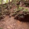 Blick auf eine im Wald liegende, rötlich braune, nach rechts ansteigende Böschung, die im oberen Teil mit Felsen durchsetzt ist. Rechts unten steht eine Schautafel.