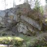 Das Bild zeigt mehrere auf einem Plateau übereinandergeschichtete Felsstücke. Nischen zwischen den Felsen sind mit Erde gefüllt oder bemoost. Auch dünne Bäume wachsen auf den Felsen. Am unteren Bildrand ist ein Wasserlauf zu sehen.