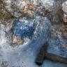Nahaufnahme von bräunlich weißem Gestein, mit blau verfärbtem Material links der Bildmitte. Ein liegender Hammer rechts dient als Größenvergleich.