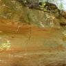 Seitlicher Blick auf die gelblich bis rötliche Wand einer aufgelassenen Sandgrube. Die Wand zeigt rinnenförmige Vertiefungen sowie oben leicht vorkragendes, lose aufliegendes Gestein.