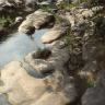 Das Bild zeigt mehrere flache Felsplatten sowie Felsblöcke in einem Flussbett. Beide Flussufer sind bewaldet, das rechte auch felsig.