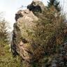 Blick auf einen steilen, oben aufgesprengten Felsenturm. Das rötlich graue Gestein ragt hinter einem mit Gebüsch bewachsenen, nach rechts aufsteigenden Felshang hervor.