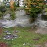 Blick auf eine halbrunde, nach rechts abgestufte Gesteinswand mit größerer Nische rechts. Die Kuppe der Wand ist mit Buschwerk und Bäumen bewachsen. Vor der Wand, auf einer Wiese, liegt links eine Feuerstelle.