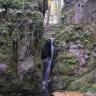 Blick auf zwei rötlich graue, stark bewachsene und bemooste Felsformationen, die am unteren Ende von einem See umschlossen sind. Etwa auf halber Höhe zwischen den Felsen stürzt sich ein schmaler Wasserfall in den See.