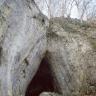 Zwischen zwei sich einander zuneigenden Felswänden befindet sich eine dreieckige Höhlenöffnung. Größere Felsstücke und abgefallenes Laub liegen vor dem Höhleneingang. Die Felswände sind oben mit Bäumen bestanden.