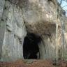 Blick auf eine hohe, rechts vorgewölbte graue Felswand. Unter der Wölbung ist eine Höhle sowie ein zweiter Eingang sichtbar. Im Vordergrund stehen drei hohe, schlanke Bäume.