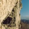 Blick auf eine steile, gelblich braune Felswand, die links entlang der Bildkante aufragt. Unterhalb der Bildmitte öffnet sich in der Felswand eine Höhle mit rautenförmigem Eingang. Rechts im Hintergrund steht auf einem Bergrücken ein Sendemast.
