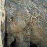 Das Bild zeigt zwei hohe, über Eck aneinanderstehende Felswände sowie einen flachen Vorplatz. Am unteren Ende der nach rechts wegführenden Felswand öffnen sich mehrere Höhleneingänge.