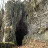 Blick auf steile, aneinander gefaltete Felswände, zwischen denen sich am Fuß eine schmale, hohe Höhle öffnet. Vor dem Eingang liegen Felsbrocken und braunes Laub.