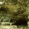 Blick auf eine nach innen gewölbte, gelblich graue Felswand. Rechts unten ist der bogenförmige Eingang zu einer Höhle erkennbar. Darüber verläuft ein fast waagerechter Riss durch das Gestein.