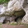 Blick auf eine weißlich graue bis braune Felswand, an deren Fuß sich Nischen und Säulen sowie rechts eine bogenförmige Höhle öffnet. Noch weiter rechts steht die Felswand etwas vor.