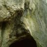 Blick auf eine steil nach links aufssteigende, ausgebleichte Felswand. Im oberen Bereich ist eine Nische, am Fuß des Gesteins, auf Bodenhöhe, eine Höhlenöffnung sichtbar.