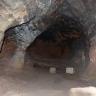 Blick in eine Höhlenkammer. Wände und Decke der Höhle bestehen aus schwärzlichem, der Eingangsbereich aus rötlichem Gestein. Zwei kleinere helle Steinbrocken liegen wie Zähne am Eingang der Höhle. 