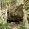 Blick auf eine steile, rechts zum Vordergrund hin abknickende Felswand. Am Fuß des bleichen Gesteins öffnete sich der flache, von zahnartigen Gesteinsbrocken gesäumte Eingang zu einer Höhle. Links stehen Bäume und wächst Gebüsch.