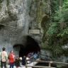 Blick auf den Eingang einer Höhle. Die Höhle liegt am unteren Ende einer hoch aufragenden Felswand. Rechts ist der Fels bewachsen. Vor dem Höhleneingang warten mehrere Menschen.