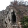 Inmitten eines auf der Kuppe sowie der rechten Seite bewachsenen Felsenhanges öffnet sich eine schmale, hohe Höhle. Eine Treppe führt hinauf zum Eingang. Auf der linken Seite der bleichen Felswand sind weitere, kleinere Höhlen und Nischen erkennbar.