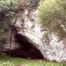 Blick auf eine bleiche, von Bäumen flankierte Felswand. In der Bildmitte, am unteren Ende der Wand, öffnet sich eine Höhle. Der nach rechts weisende, verschattete Eingang ähnelt im Umriss einer Pfeilspitze.