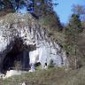 Das Bild zeigt einen hügelartig geformten, dreieckigen Felsen, dessen Kuppe mit Bäumen und Sträuchern bewachsen ist. Das bemooste, weißlich graue Gestein weist an der Vorderseite einen hohen und breiten Höhleneingang auf.