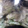 Blick auf eine hohe Felsenkammer, in der sich unten eine breite, flache Höhle und oben ein Ausstieg öffnet, durch den Licht hereinkommt.