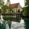 Eine schlichte, mit einem spitzen Turm versehene Kirche spiegelt sich in einer unmittelbar angrenzenden Wasserfläche.