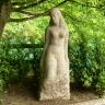 Auf dem Bild sieht man die aus einem hellen Steinblock modellierte Skulptur einer Frau. Die Skulptur steht im Schatten eines Baumes und ist von Sträuchern umgeben.