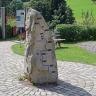 Blick auf einen sich nach oben verjüngenden Steinblock. Auf einer Seite ist der Lauf der Donau eingraviert. Die Stele steht auf einem gepflasterten Weg. Im Hintergrund sind Hinweistafeln aufgestellt.