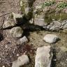 Blick von oben auf einen kleinen Quellaustritt. Das obere Ufer des Wasserlaufs ist von einer niedrigen Steinmauer gesäumt. Links und nach unten besteht das Ufer aus losen Steinen und flachen Stellen.