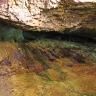 Aufwärts gerichteter Blick auf eine quer durchs Bild verlaufende Spalte zwischen Gesteinswänden. Der Bereich um die Spalte ist grünlich verfärbt, zum Vordergrund hin folgen rostbraune Farben.