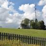 Im Mittelpunkt des Bildes steht ein kleiner gemauerter Pavillon mit Überdachung. Der Aussichtspunkt ist auf einer begrünten, flachen Bergkuppe. Ein paar Bäume und ein Sendemast stehen rechts vom Pavillon. Im Vordergrund verläuft ein Holzzaun.