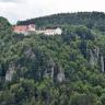 Blick auf einen bewaldeten Bergkegel. An den Seiten des Berges treten mehrere Felszacken hervor. Auf der flachen Kuppe des Berges stehen mehrere Gebäude, die miteinander verbunden sind.