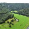 Blick aus großer Höhe auf eine Schlinge der Donau. Der Fluss verläuft von rechts unten zur Bildmitte hin und verschwindet dort zwischen bewaldeten Bergen. Das linke Ufer ist flach, daran schließen sich bewaldete Berge an, die bis zum Hintergrund reichen. 