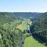 Blick aus großer Höhe über ein langgezogenes Tal mit Flusslauf. Das Tal ist hufeisenförmig von bewaldeten Bergen eingerahmt. Am Berghang links im Vordergrund stehen bleiche Felsen heraus.