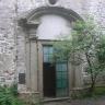 Blick auf ein steinernes, mit rundem Steindach versehenes Eingangsportal. Das Portal befindet sich in einer Fassade aus Mauersteinen. Die linke Hälfte der geteilten Eingangstür steht offen. Rechts daneben wächst ein kleiner Baum.