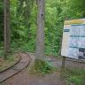 Das Foto zeigt ein abzweigendes Gleis in einem Wald. Das eine Gleis verläuft gerade, das andere in einem Bogen. Rechts steht zudem eine große Informationstafel mit Wanderkarten rund um die Gemeinde Spiegelberg.
