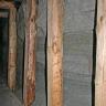 Das Bild zeigt eine graue Stollenwand in einem alten Bergwerk. Die Stollenwand wird durch mehrere Holzbalken gestützt. Oben links ist die durch Querbalken gestützte Stollendecke zu erkennen.